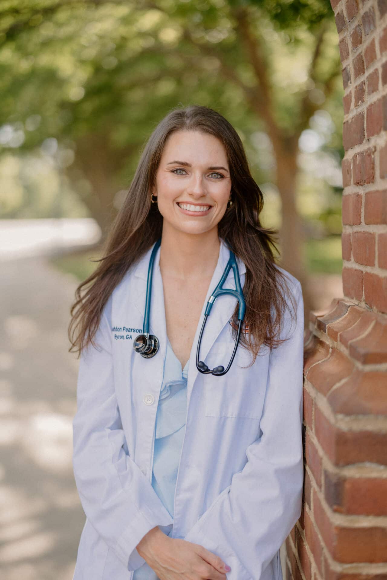 A woman in a white coat with a stethoscope smiles, standing in front of a brick wall with trees in the background.